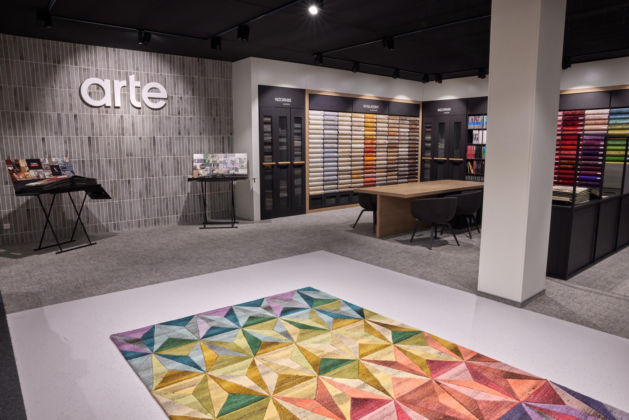 Salon z ekspozycją próbek podłogowych, wzorników i dywanu z geometrycznym wzorem w odcieniach zieleni, żółci, różu i fioletu na pierwszym planie. W tle widoczne regały z próbkami i logo 'arte'...