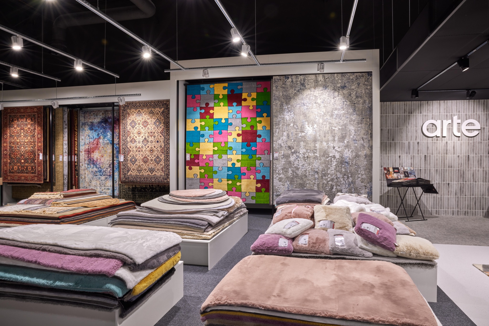 Wystawa dywanów w sklepie: różnorodne wzory, od tradycyjnych po nowoczesne, w tym dywan-puzzle, ułożone na białych podestach pod oświetleniem szynowym, z logo 'arte' na tle ściany z płytek.