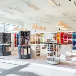 Salon z ekspozycją próbek dywanów i wykładzin w różnych kolorach i wzorach, oświetlony wiszącymi lampami i halogenami