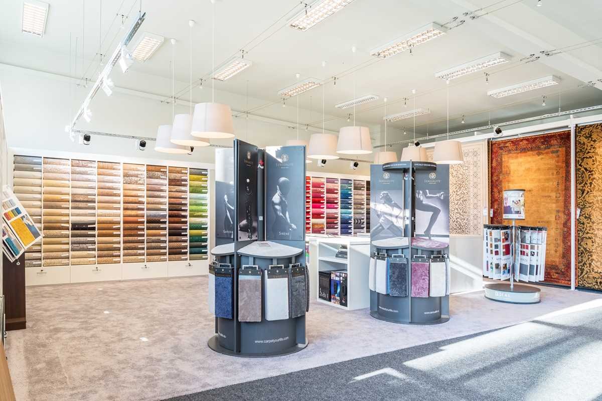 Salon z ekspozycją różnorodnych próbek podłogowych, w tym wykładzin dywanowych zwiniętych w rolki i wiszących próbek tkanin, oświetlony wiszącymi lampami z abażurami.