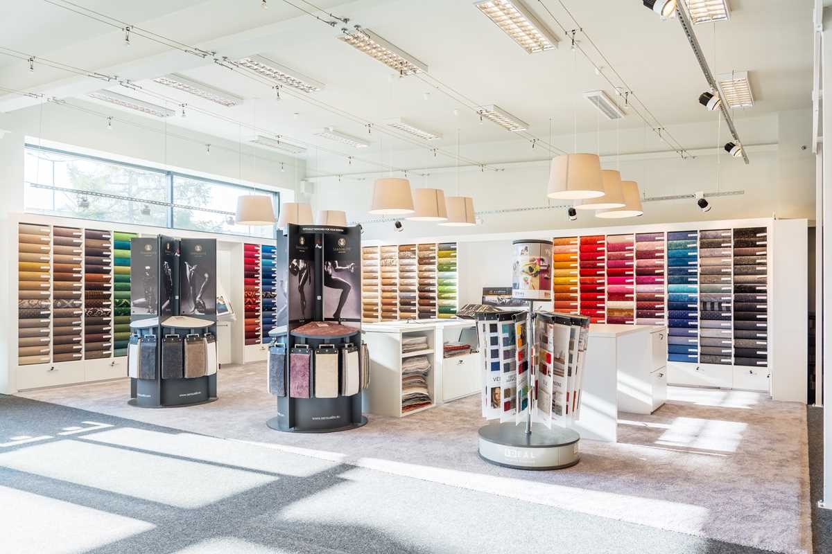 Salon z ekspozycją próbek dywanów i wykładzin w różnych kolorach i wzorach, oświetlony wiszącymi lampami i halogenami