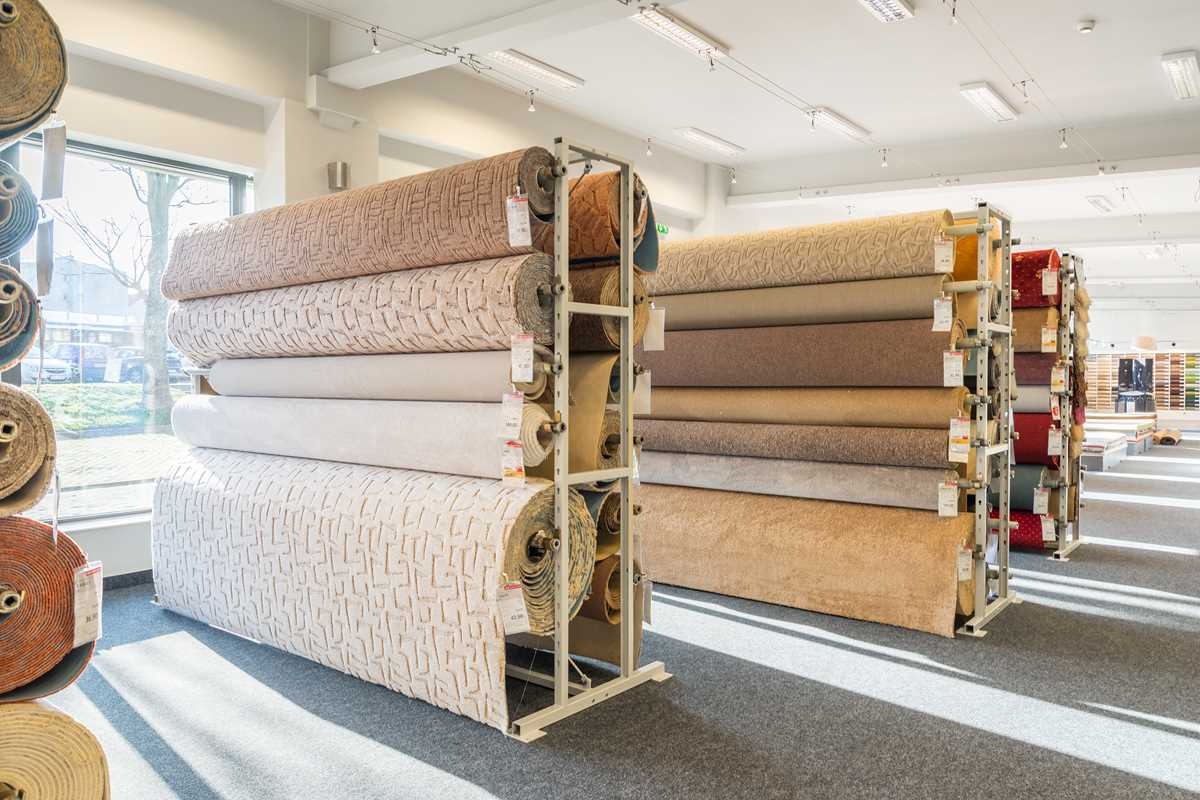 Rzędy rolek z różnymi wzorami i odcieniami beżu, brązu i kremu, ustawione na metalowych regałach w dobrze oświetlonym sklepie z szarą wykładziną podłogową.