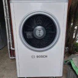 Biała pompa ciepła Bosch zamontowana na zewnątrz budynku, widoczne rury w izolacji i fragment otoczenia z kostki brukowej.