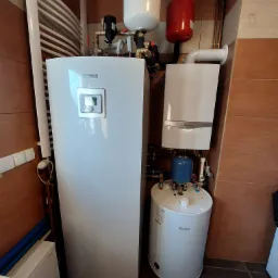 Kotłownia z dwoma zbiornikami: pionowy biały Bosch z panelem sterowania i mniejszy, również biały, marki Vaillant, z niebieskim zbiornikiem wyrównawczym, na tle płytek imitujących drewno.