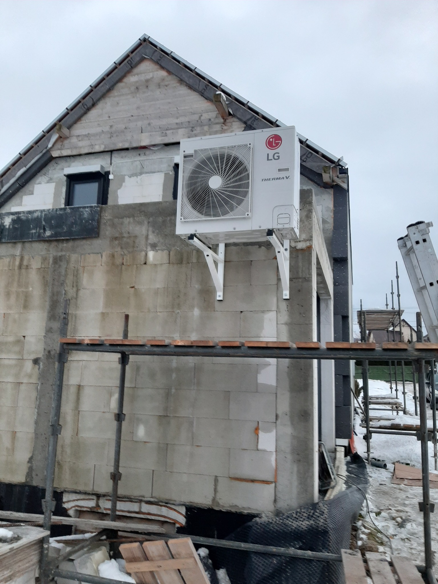 Biała zewnętrzna jednostka pompy ciepła LG Therma V zamontowana na ścianie budynku w trakcie budowy, widoczne rusztowania i materiały budowlane.