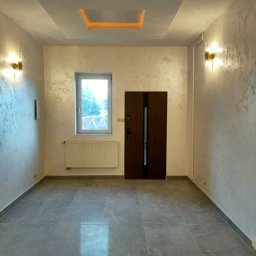 Yuriy Romanenko usługi budowlane - Wykończone wnętrze pokoju z szarymi płytkami podłogowymi, ścianami z dekoracyjną fakturą, oknem, grzejnikiem oraz podwieszanym sufitem z oświetleniem LED.
