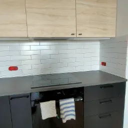 Kuchnia w trakcie remontu: ułożone białe płytki typu metro, widoczne puszki elektryczne do podłączenia, czarne szafki dolne, jasne szafki górne z drewnopodobnym dekorem, płyta indukcyjna.