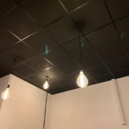 Ciemny sufit kasetonowy z trzema wiszącymi żarówkami w stylu retro, widoczne haczyki montażowe, białe ściany w tle.