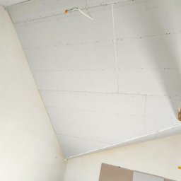 Yuriy Romanenko usługi budowlane - Montaż płyt kartonowo-gipsowych na skośnym suficie poddasza, widoczne łączenia i wkręty, przewody elektryczne wystające z sufitu, materiały budowlane i profile na podłodze.