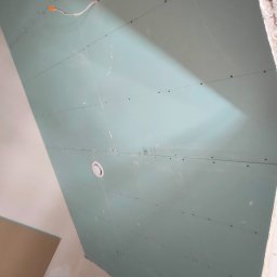 Yuriy Romanenko usługi budowlane - Niebieska płyta karton gips zamontowana na suficie, widoczne wkręty, otwór na oświetlenie i wystający kabel elektryczny.