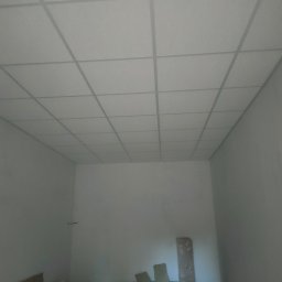 Yuriy Romanenko usługi budowlane - Kwadratowa podwieszana konstrukcja sufitowa z widocznymi elementami montażowymi, częściowo ukończona, z materiałami budowlanymi i odpadami na podłodze w pustym pomieszczeniu.