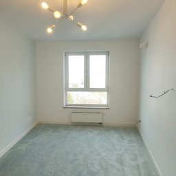 Yuriy Romanenko usługi budowlane - Pomalowane na biało puste pomieszczenie z turkusową wykładziną dywanową, oświetlone nowoczesną lampą sufitową z wieloma żarówkami, okno z grzejnikiem pod spodem.