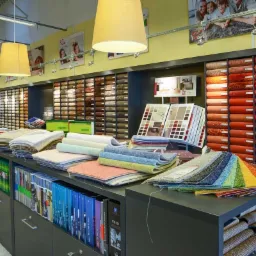 Ekspozycja próbek podłogowych w sklepie: rolki i kwadraty dywanowe w odcieniach beżu, brązu, czerwieni i tęczy, ułożone na półkach i blacie szafki z katalogami.