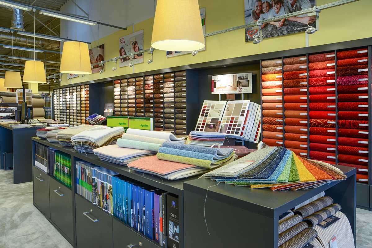 Ekspozycja próbek podłogowych w sklepie: rolki i kwadraty dywanowe w odcieniach beżu, brązu, czerwieni i tęczy, ułożone na półkach i blacie szafki z katalogami.