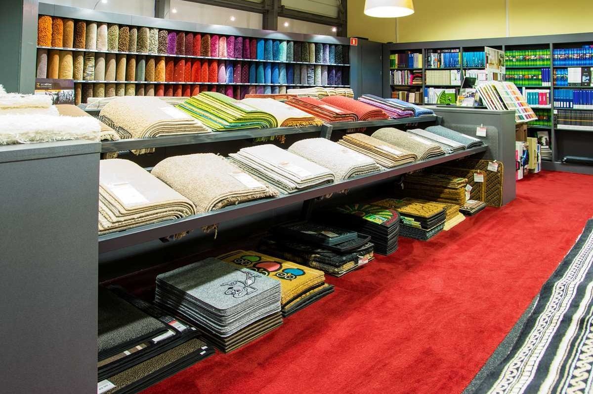 Ekspozycja różnorodnych wykładzin i dywanów w sklepie: próbki ułożone na półkach, widoczne wzory, faktury i kolory, czerwona wykładzina podłogowa.