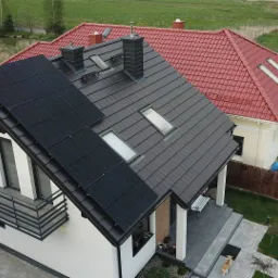 Widok z góry na dom jednorodzinny z panelami słonecznymi na dachu, czarna dachówka, balkon z metalową balustradą, obok dom z czerwoną dachówką, w tle łąka i staw.