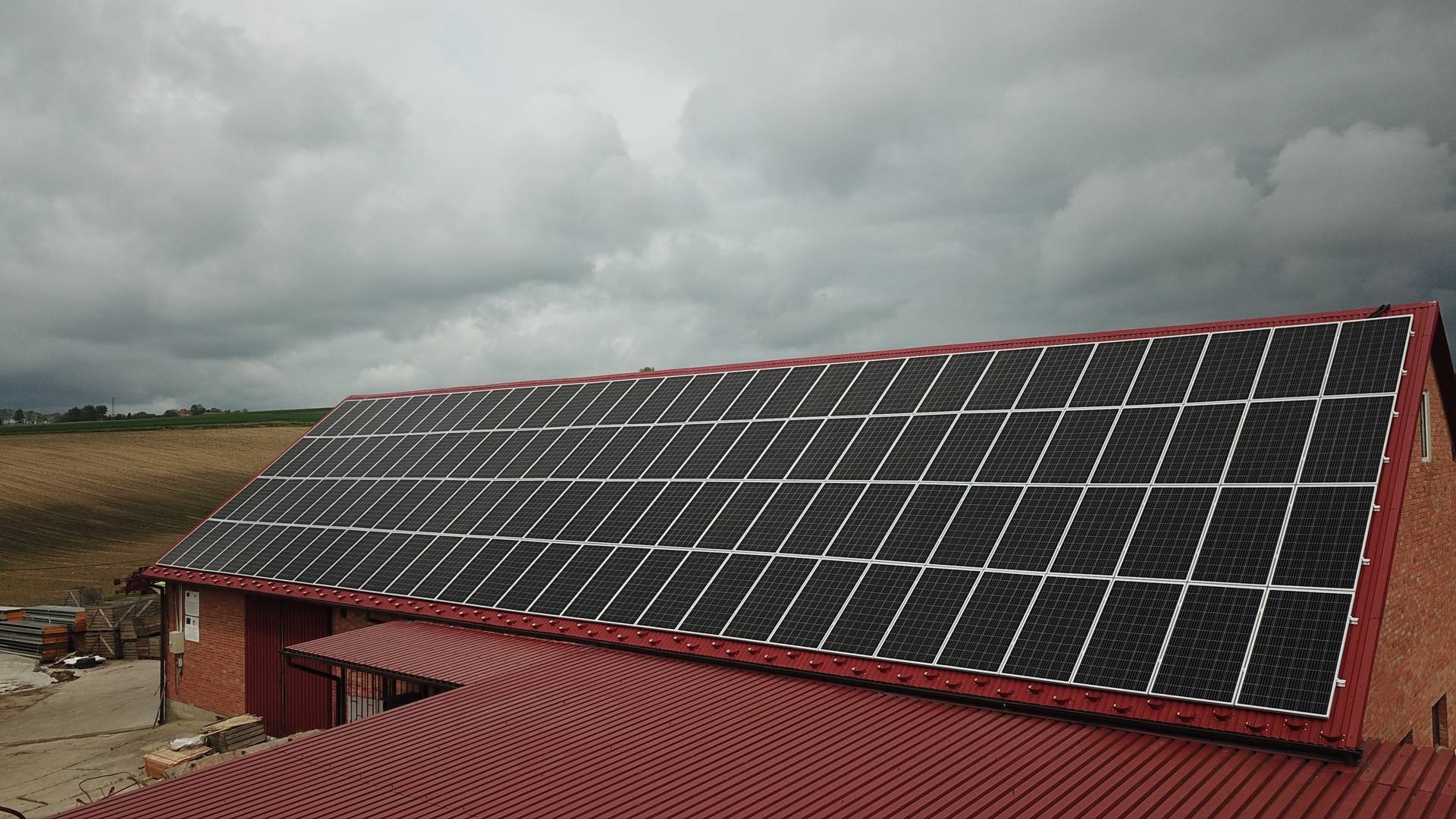 Instalacja fotowoltaiczna na dachu budynku gospodarczego z czerwonej blachy trapezowej, widok z góry na tle pochmurnego nieba i pól uprawnych.