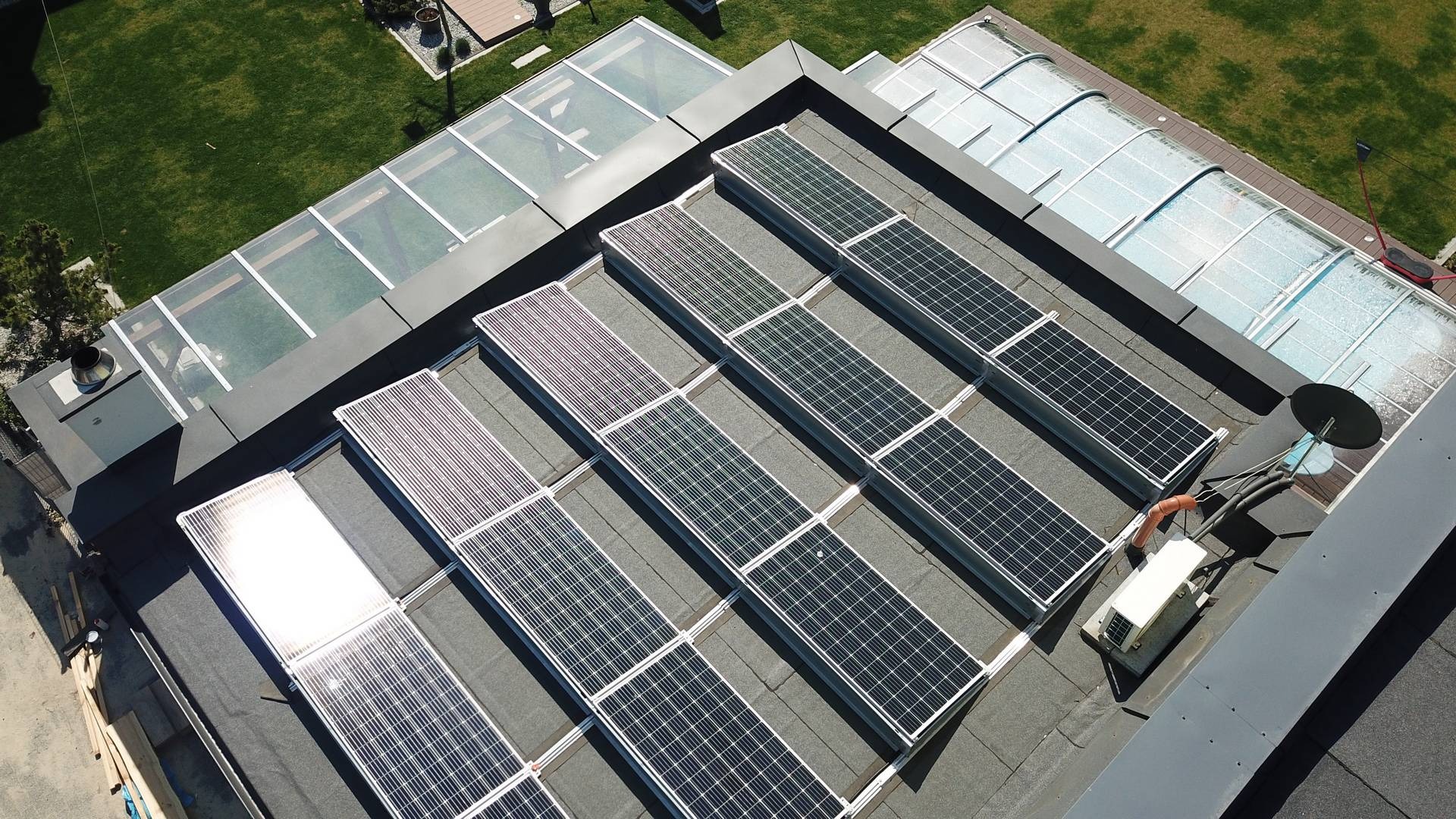 Widok z góry na panele fotowoltaiczne zamontowane na dachu budynku z basenem i przeszklonym tarasem, widoczna jednostka zewnętrzna klimatyzacji i elementy instalacji.