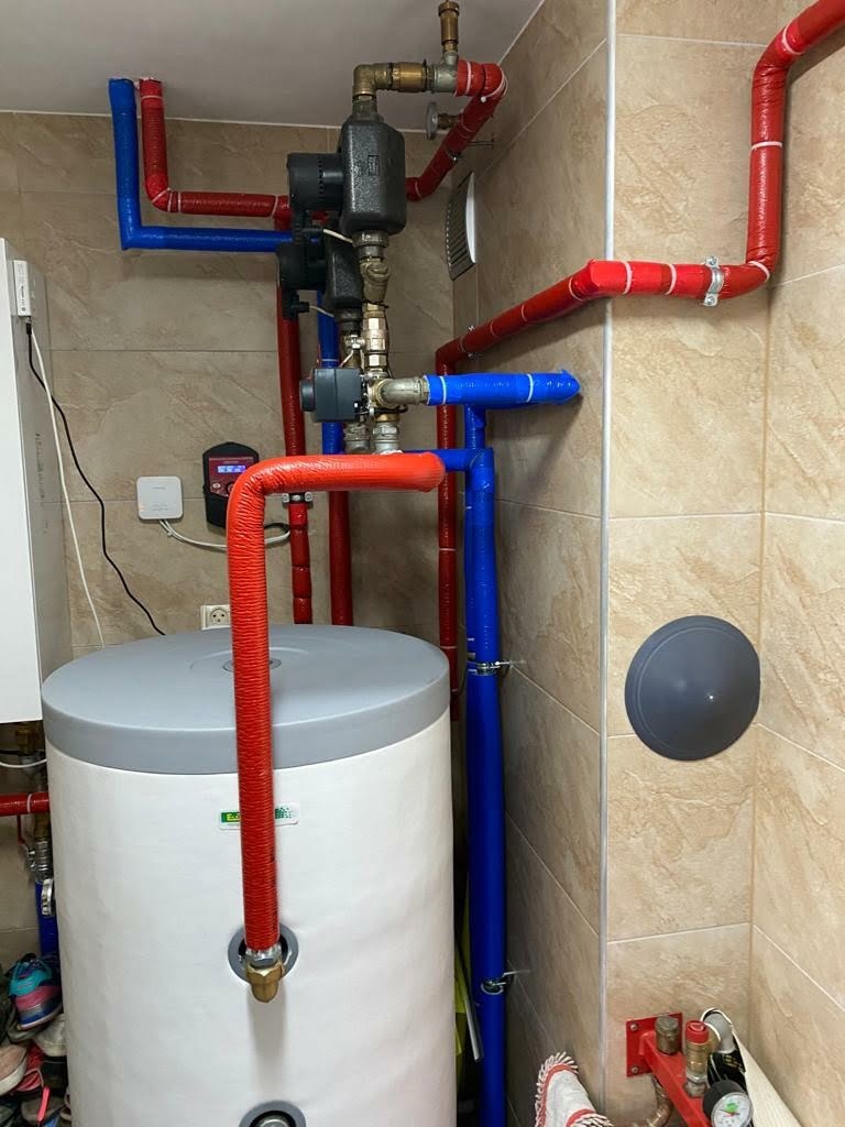 Instalacja grzewcza z białym zasobnikiem ciepłej wody, rurami w izolacji czerwonej i niebieskiej, oraz elementami armatury na tle beżowych płytek ceramicznych.