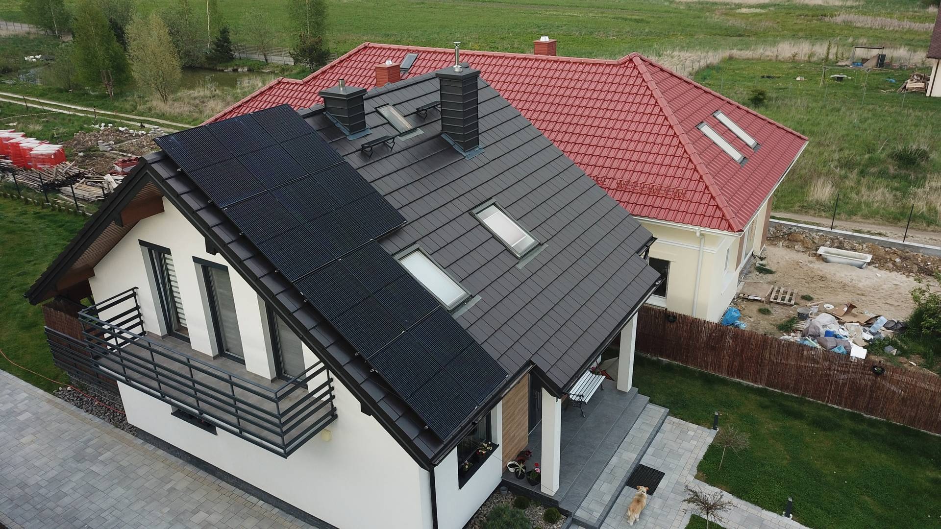 Widok z góry na dom jednorodzinny z panelami słonecznymi na dachu, czarna dachówka, balkon z metalową balustradą, obok dom z czerwoną dachówką, w tle łąka i staw.