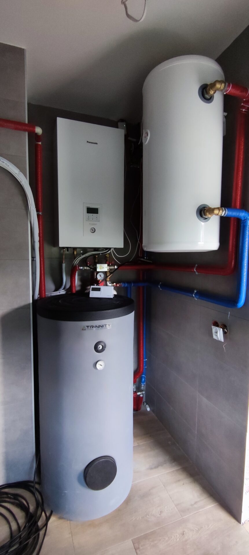 Zestaw urządzeń grzewczych: szary zbiornik Trinity, biały bojler i wiszący panel Panasonic z wyświetlaczem, połączone rurami w kolorach czerwonym i niebieskim, zamontowane na ścianie z szarych płytek.