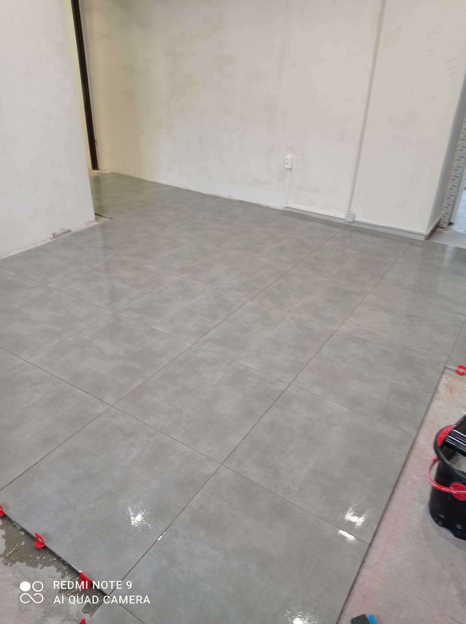 Ułożone szare płytki podłogowe w pustym pomieszczeniu, widoczne krzyżyki dystansowe i wiadro z narzędziami do układania płytek.