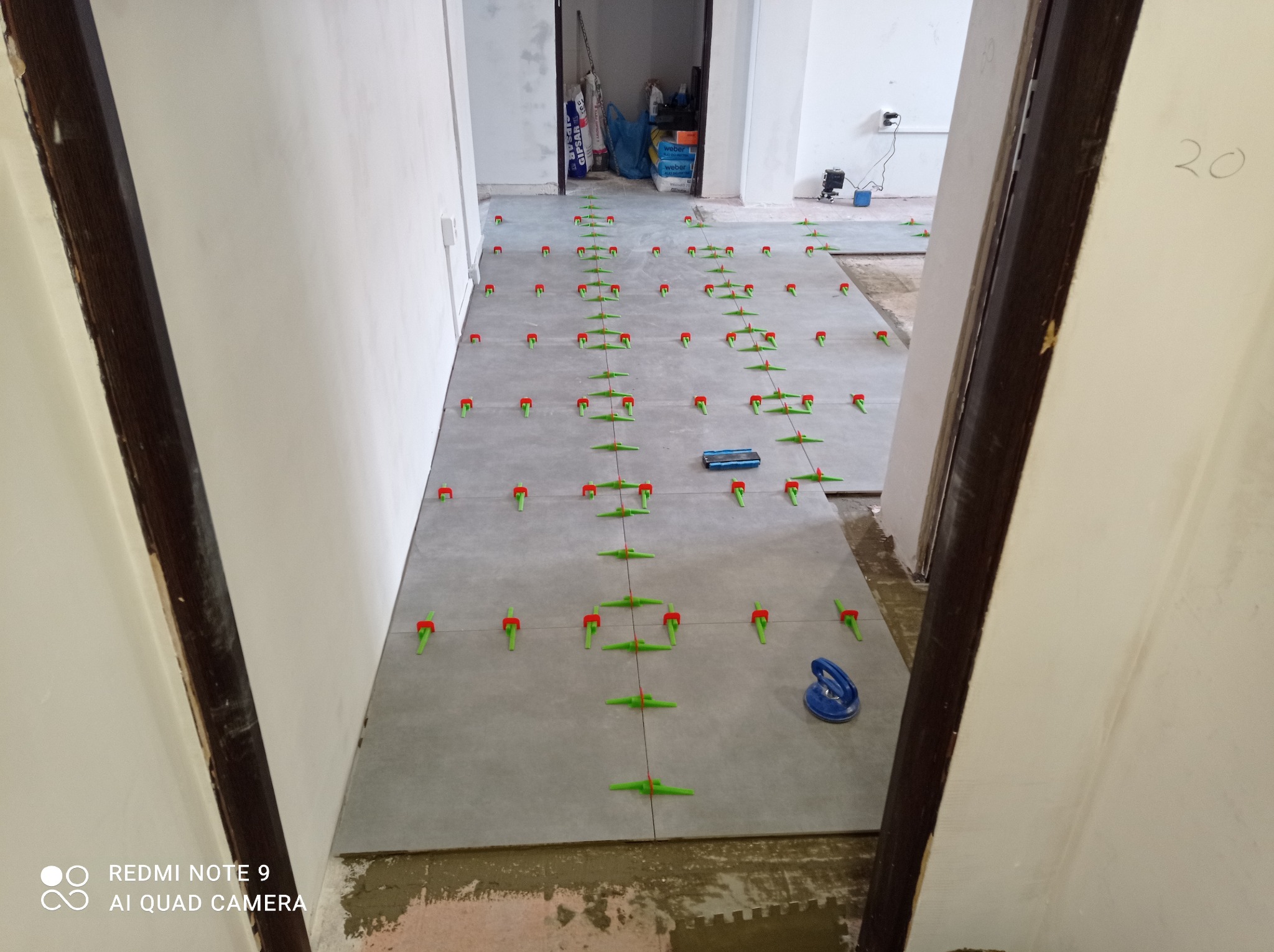 Szare płytki podłogowe w trakcie układania, z użyciem systemów poziomowania płytek (czerwone i zielone kliny), widoczne narzędzia i worki z materiałami budowlanymi w tle.