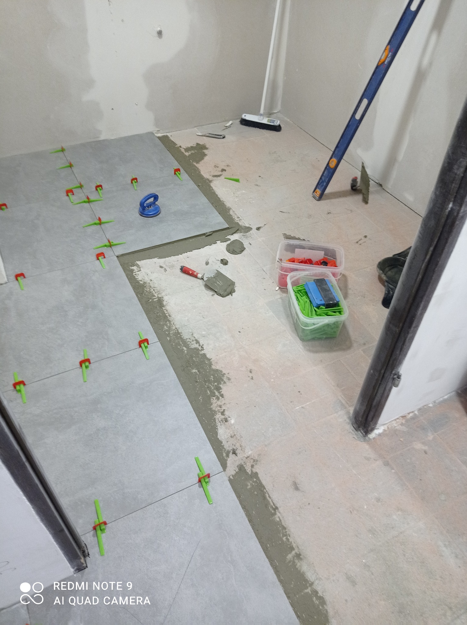 Układanie szarych płytek podłogowych w trakcie remontu łazienki, widoczne poziomica, przyssawka do płytek i system poziomowania płytek.