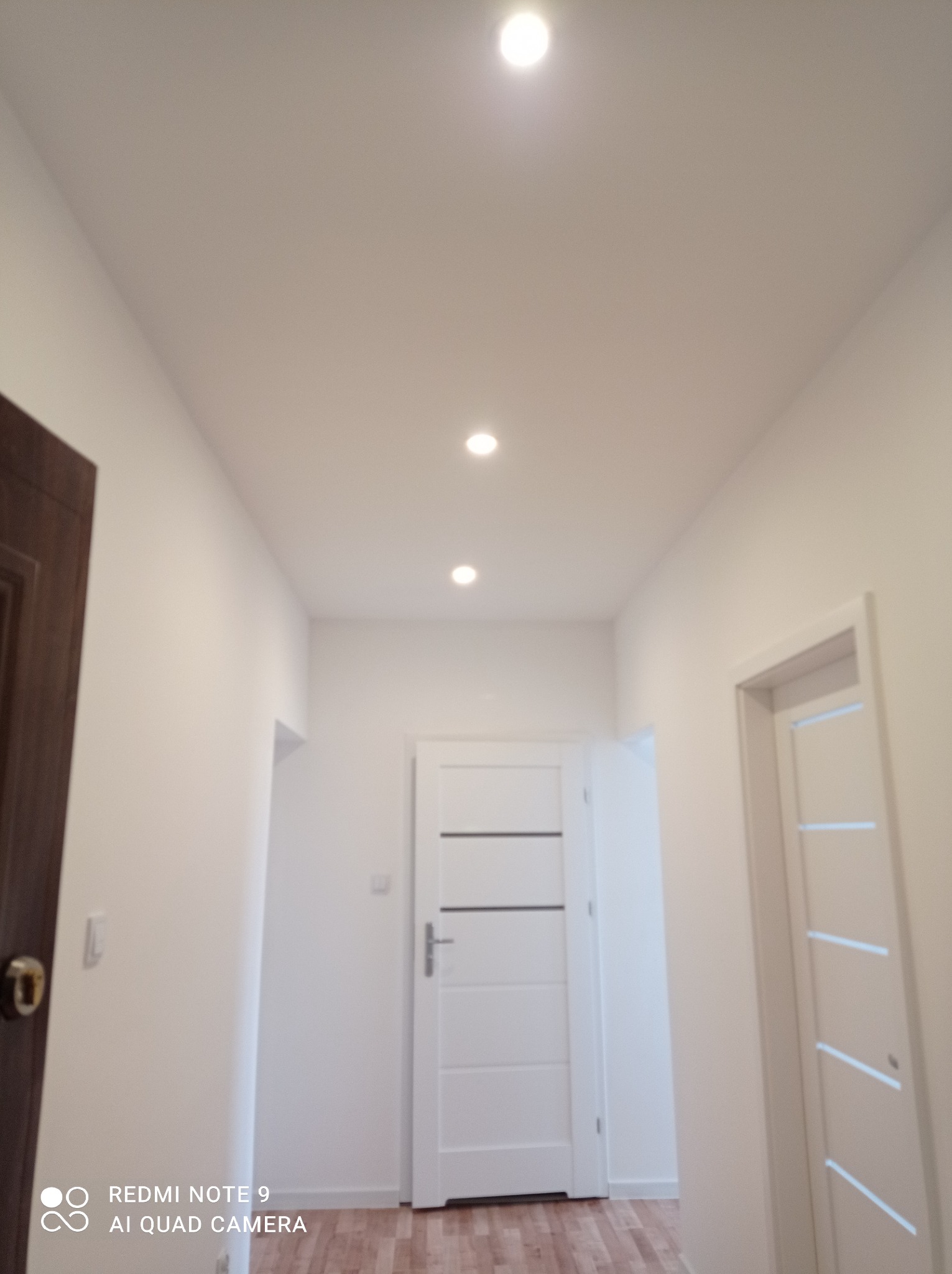 Korytarz po remoncie z trzema parami drzwi, oświetleniem punktowym w suficie i podłogą imitującą drewno.