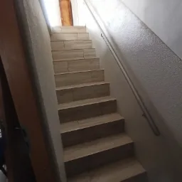 Schody wyłożone płytkami imitującymi drewno, z metalową poręczą na ścianie o szorstkiej fakturze, prowadzące do drzwi w górnej części kadru.