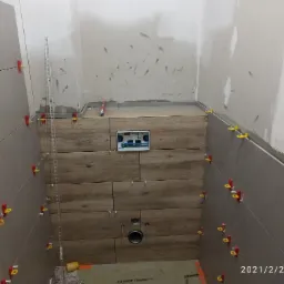 Świeżo ułożone, szare płytki ścienne z czerwono-żółtymi krzyżykami dystansowymi, kontrastujące z płytkami imitującymi drewno na zabudowie stelaża podtynkowego WC; ściany w surowym stanie przed...