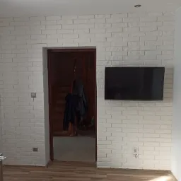 Ściana wyłożona białymi cegłami dekoracyjnymi, z zamontowanym telewizorem i wejściem do innego pomieszczenia z drewnianymi schodami.