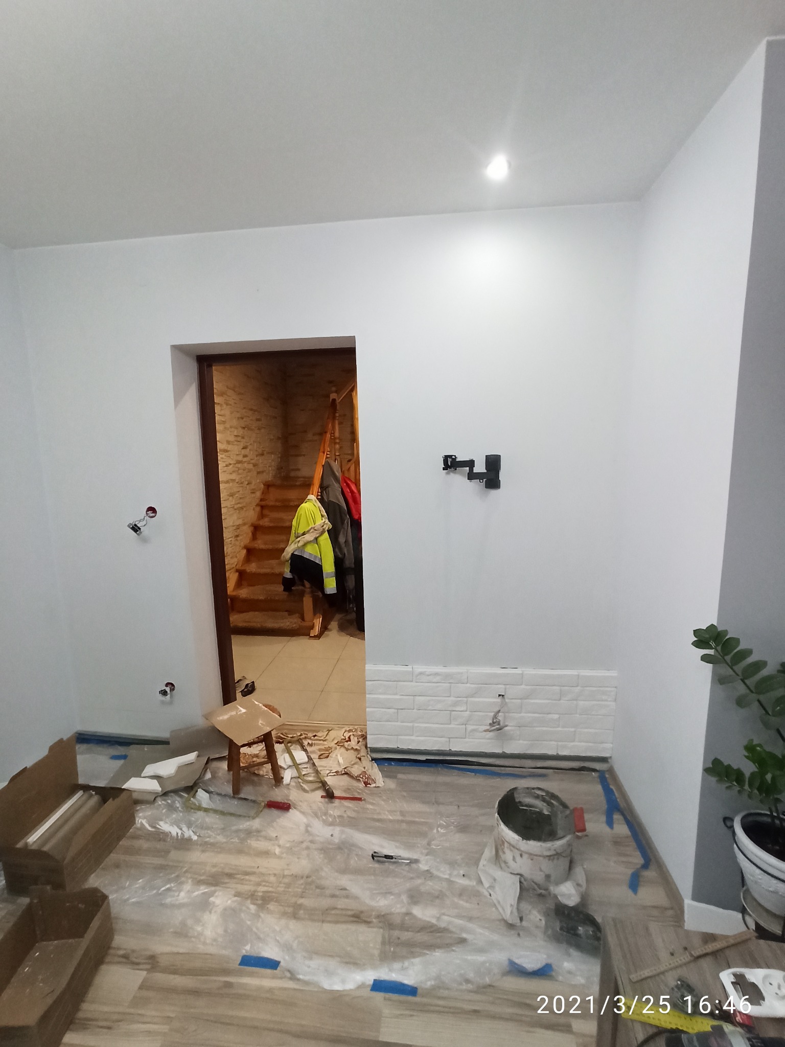 Wnętrze w trakcie prac wykończeniowych: częściowo położona biała cegiełka na ścianie, zabezpieczona podłoga folią, widoczne narzędzia malarskie i kartony, w tle drewniane schody i wiszące ubrania.