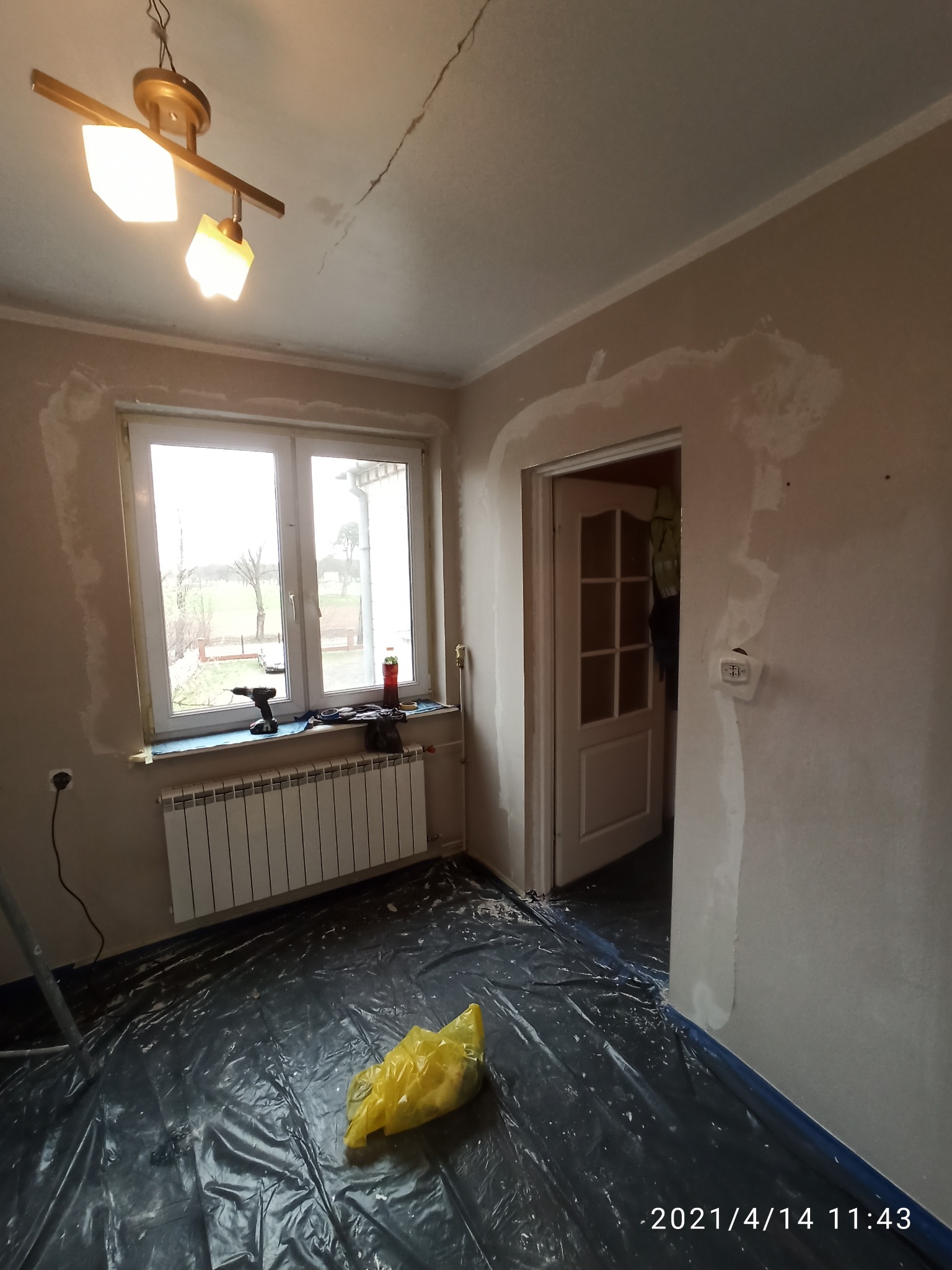 Pokój w trakcie przygotowania do prac wykończeniowych: zabezpieczona folią podłoga, szpachlowane ściany i okolice okna, widoczny sprzęt na parapecie oraz pęknięcie na suficie.