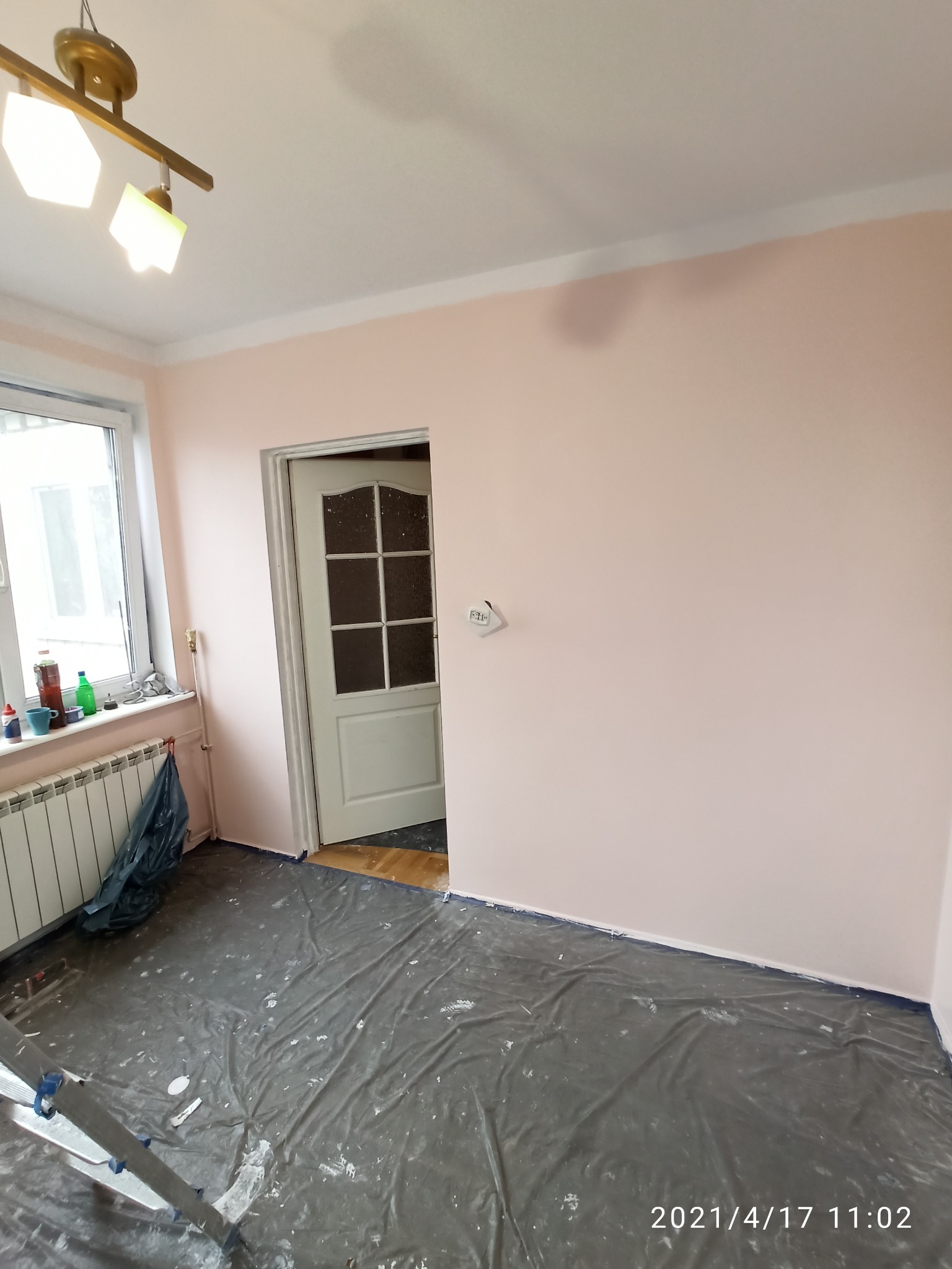 Świeżo pomalowane na różowo ściany w pokoju, zabezpieczona folią podłoga, widoczny fragment drabiny malarskiej, okno z grzejnikiem oraz drzwi.