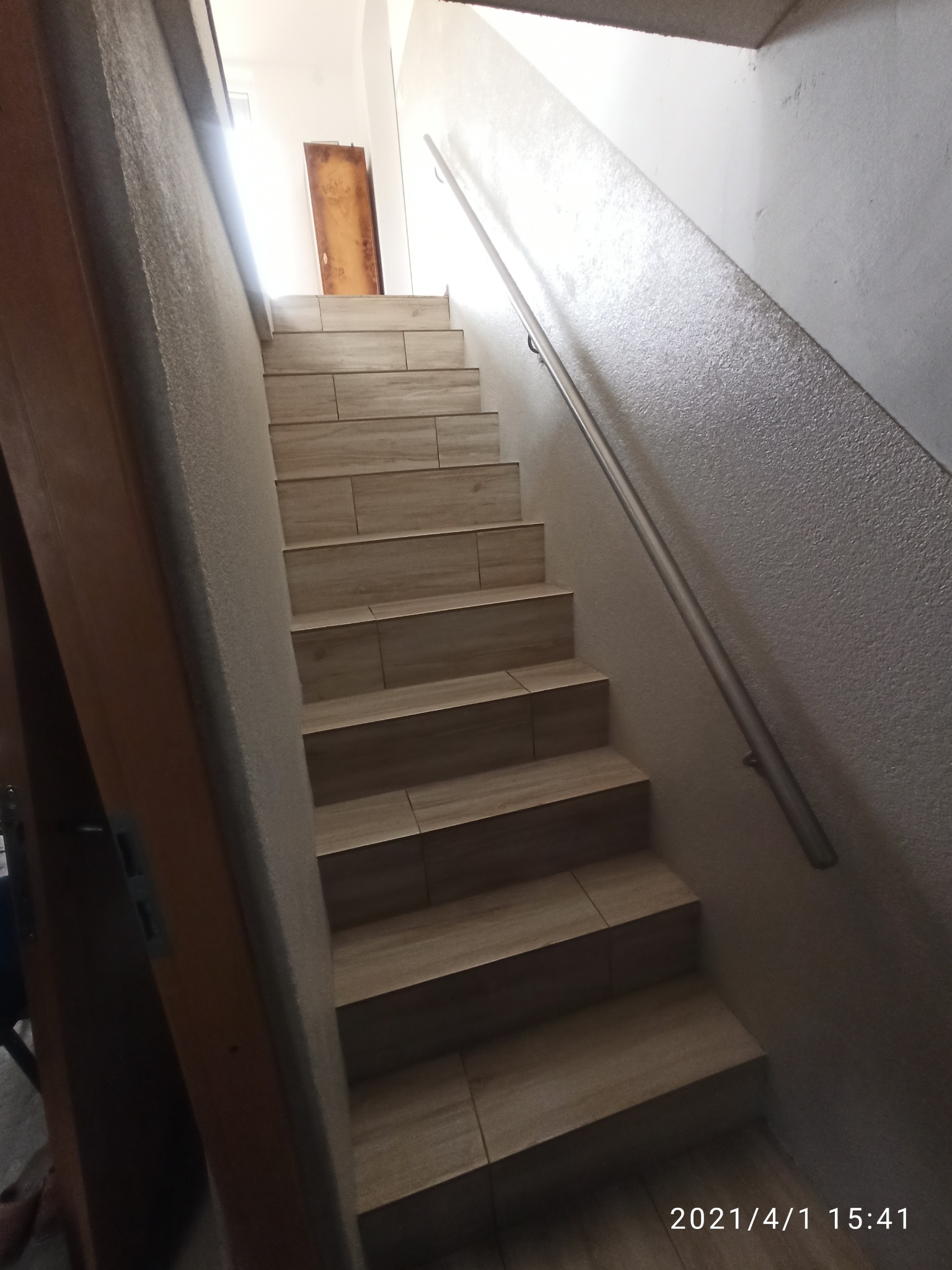 Schody wyłożone płytkami imitującymi drewno, z metalową poręczą na ścianie o szorstkiej fakturze, prowadzące do drzwi w górnej części kadru.