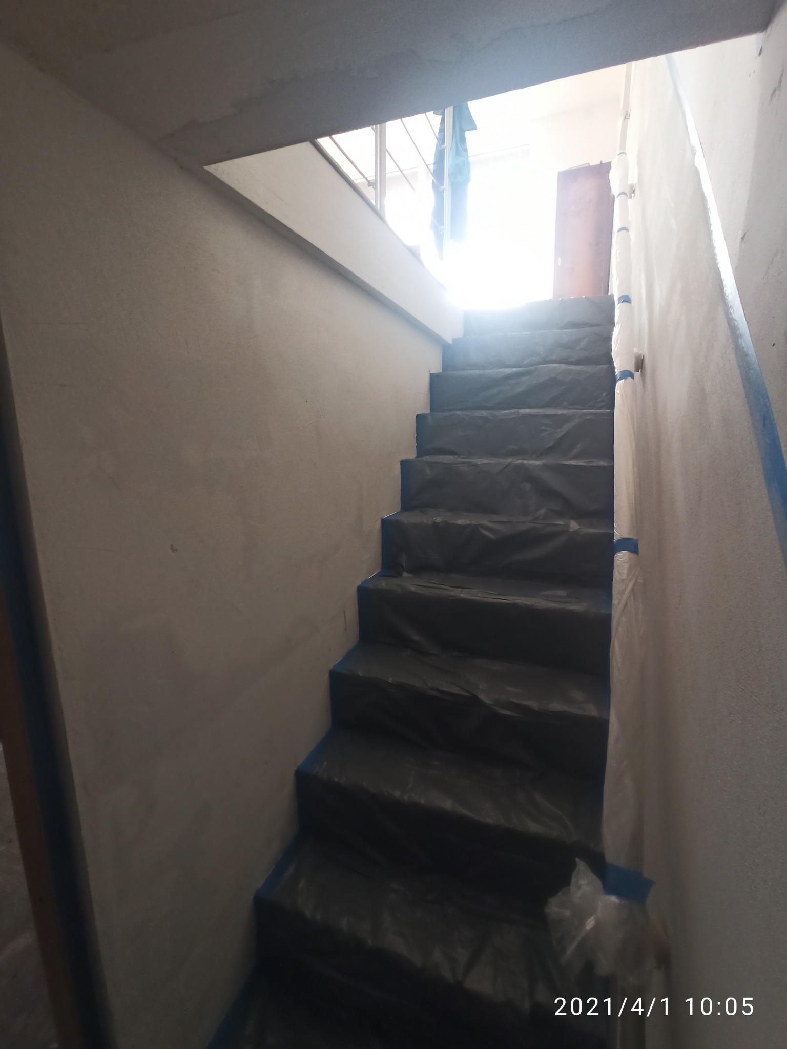 Schody zabezpieczone folią malarską i taśmą przed malowaniem ścian; widoczne świeżo otynkowane ściany i jasne światło wpadające z góry.