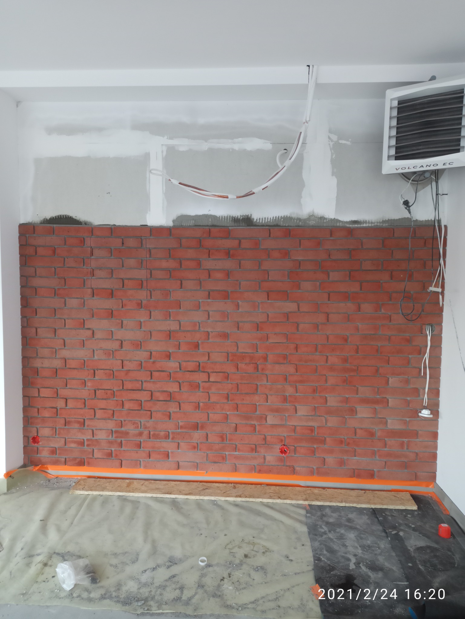 Ściana z czerwonej cegły dekoracyjnej w trakcie montażu, widoczne przewody elektryczne, osłonięta podłoga i grzejnik kanałowy Volcano EC.