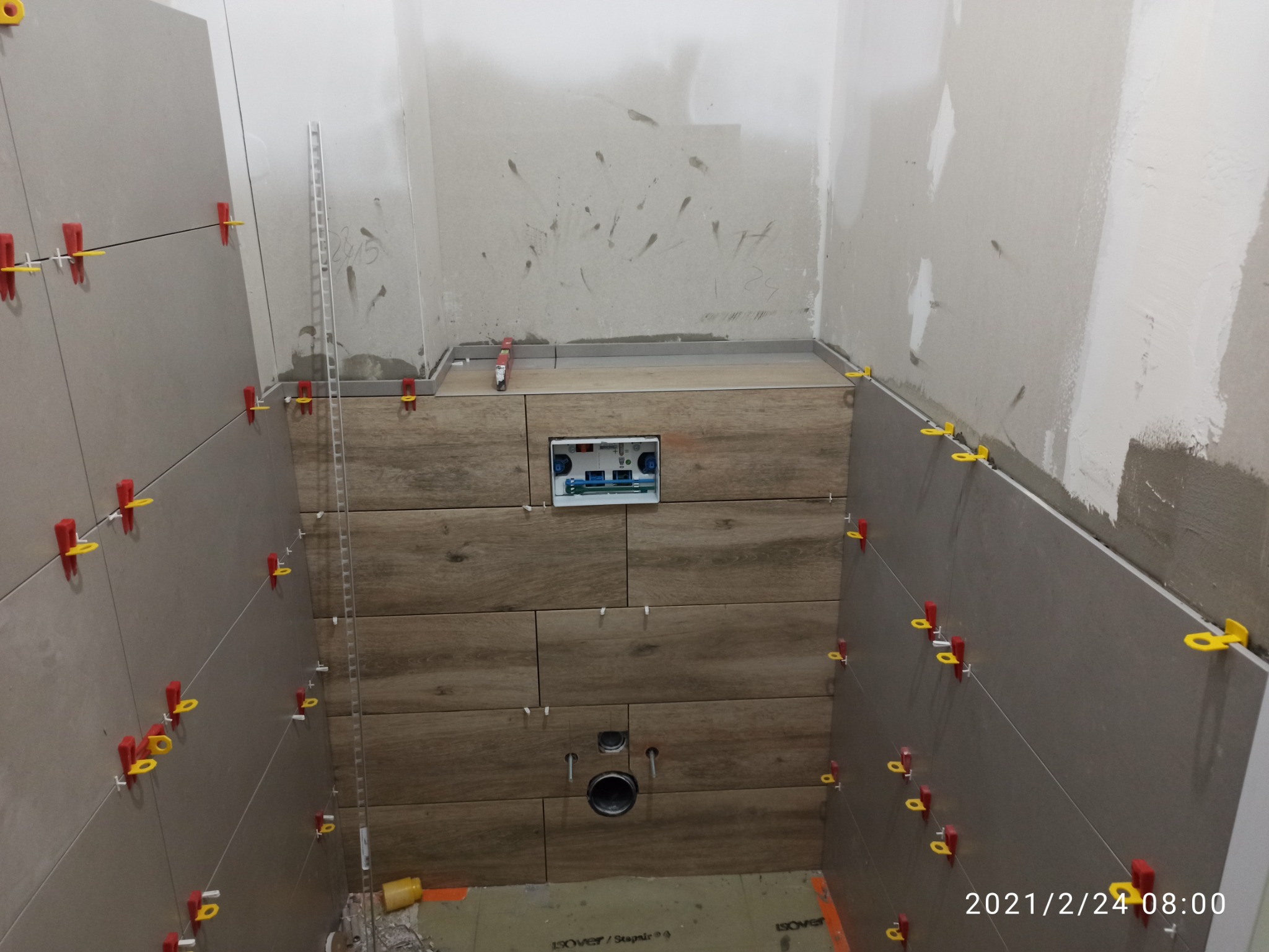 Świeżo ułożone, szare płytki ścienne z czerwono-żółtymi krzyżykami dystansowymi, kontrastujące z płytkami imitującymi drewno na zabudowie stelaża podtynkowego WC; ściany w surowym stanie przed...