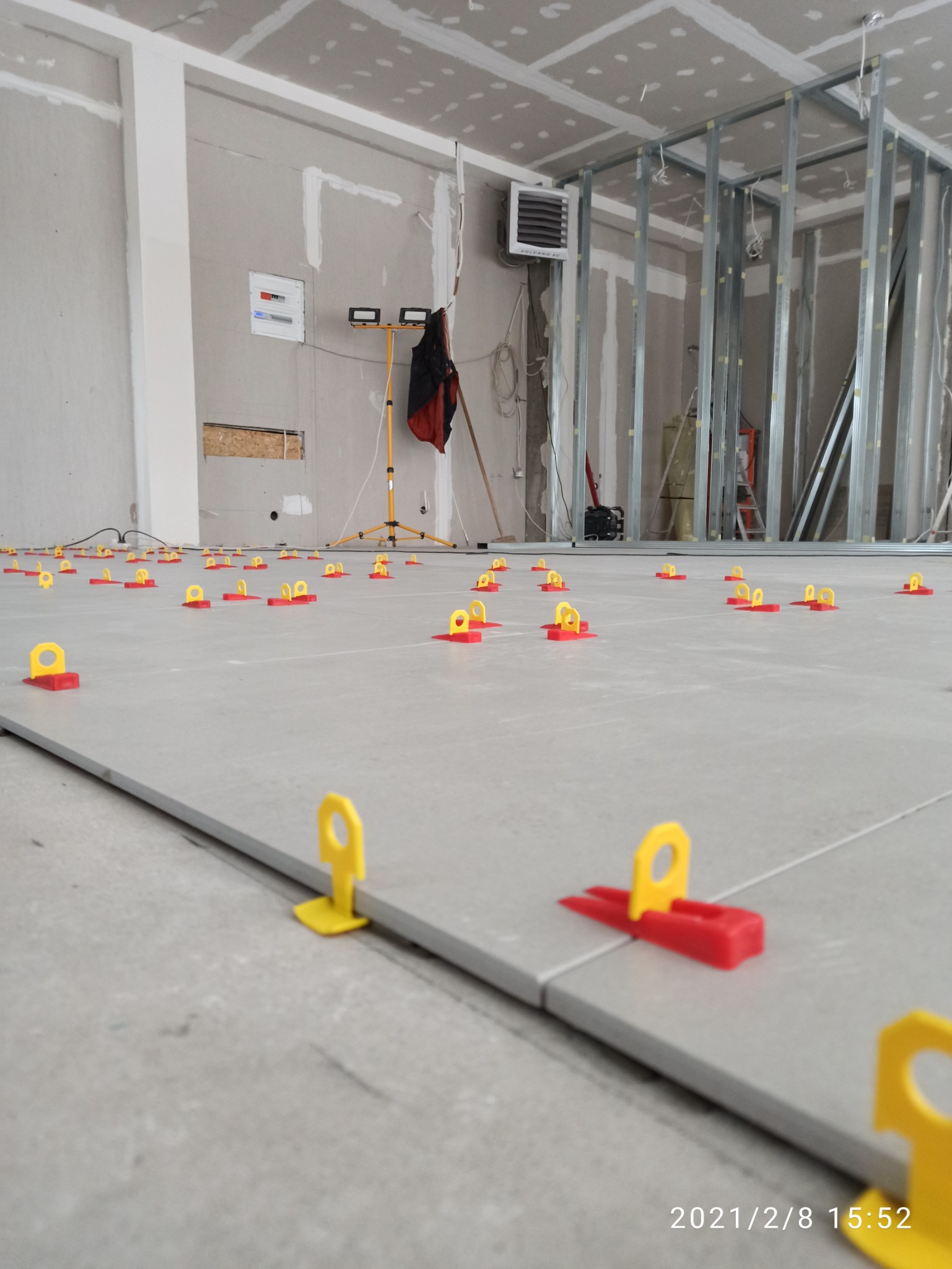 Szare płytki podłogowe w trakcie układania, z użyciem czerwono-żółtych klipsów poziomujących, widoczne metalowe profile ścian działowych w tle.