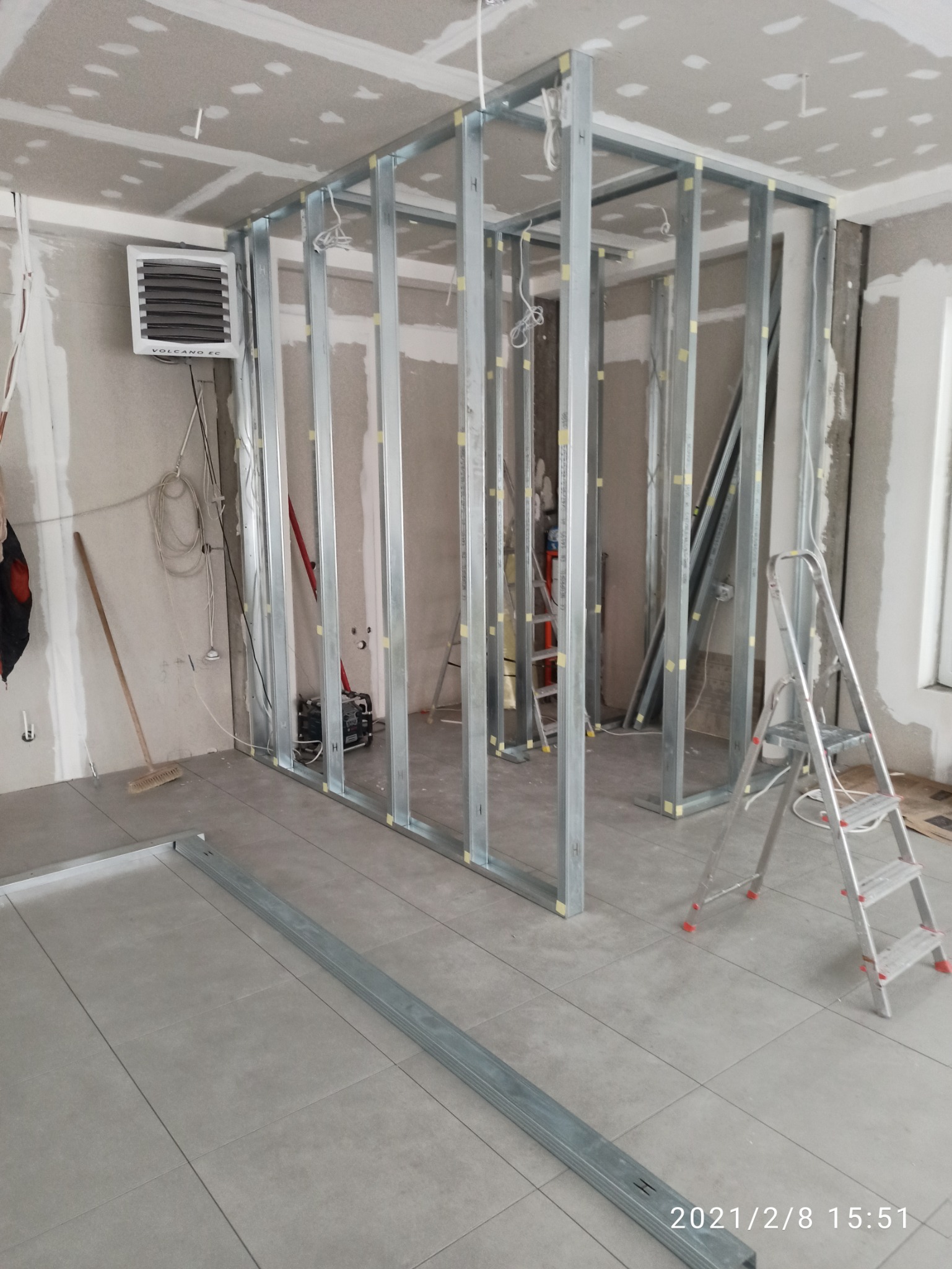Metalowa konstrukcja szkieletowa ścianki działowej w trakcie montażu w pomieszczeniu z wykończoną podłogą z szarych płytek, widoczna drabina, grzejnik ścienny Volcano i materiały budowlane.