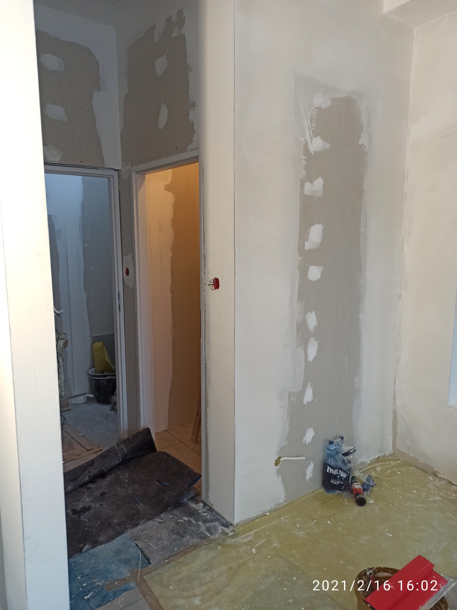 Wnętrze w trakcie prac przygotowawczych przed malowaniem: ściany z widocznymi łatami gipsowymi, zabezpieczona podłoga, butelki z wodą i narzędzia malarskie w rogu pomieszczenia.