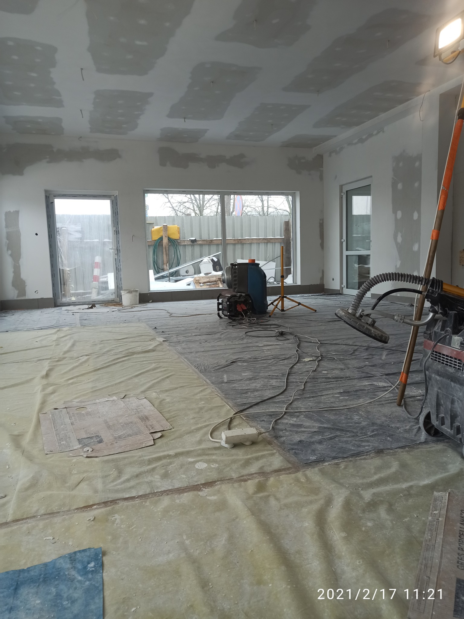 Wnętrze w trakcie prac wykończeniowych: widoczne płyty gipsowo-kartonowe na suficie z widocznymi wkrętami, zakryta folią podłoga, szlifierka do gładzi na podłodze i lampa halogenowa na statywie.