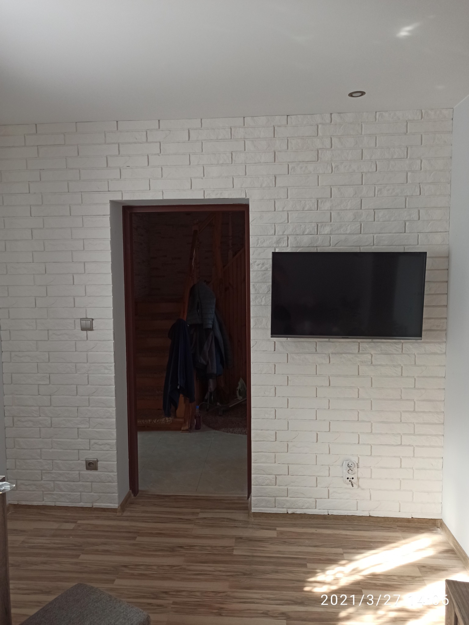 Ściana wyłożona białymi cegłami dekoracyjnymi, z zamontowanym telewizorem i wejściem do innego pomieszczenia z drewnianymi schodami.