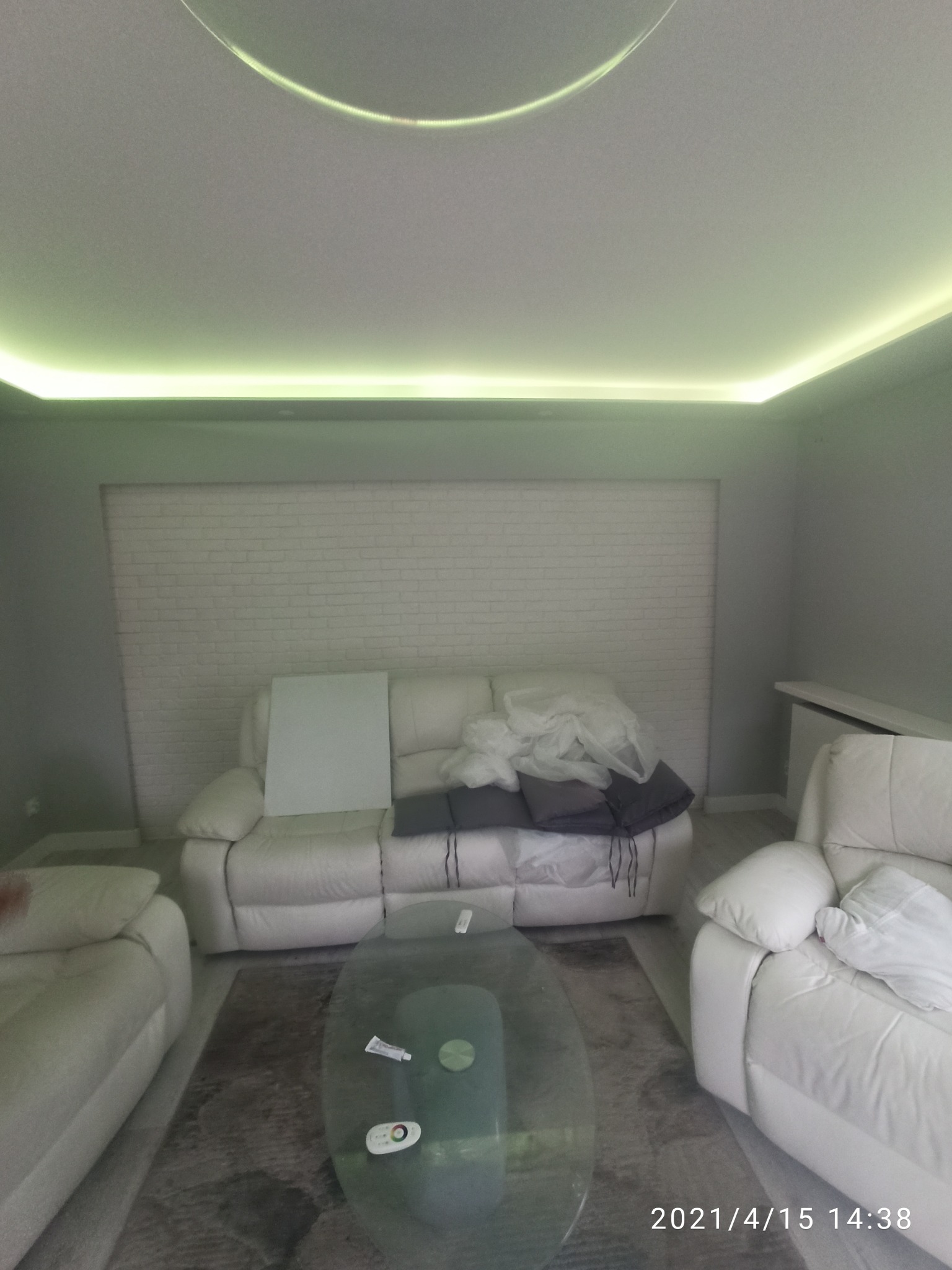 Salon z białymi skórzanymi sofami, szklanym stolikiem i oświetleniem LED pod sufitem. Ściana z imitacji białej cegły. Na sofie leżą poduszki i folia ochronna.