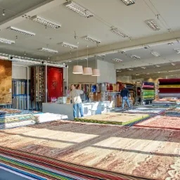 Wnętrze sklepu z szerokim wyborem dywanów i wykładzin, prezentujących różnorodne wzory, kolory i faktury. Klienci przy ladzie obsługiwani przez personel. Widoczne oświetlenie sufitowe i regały...