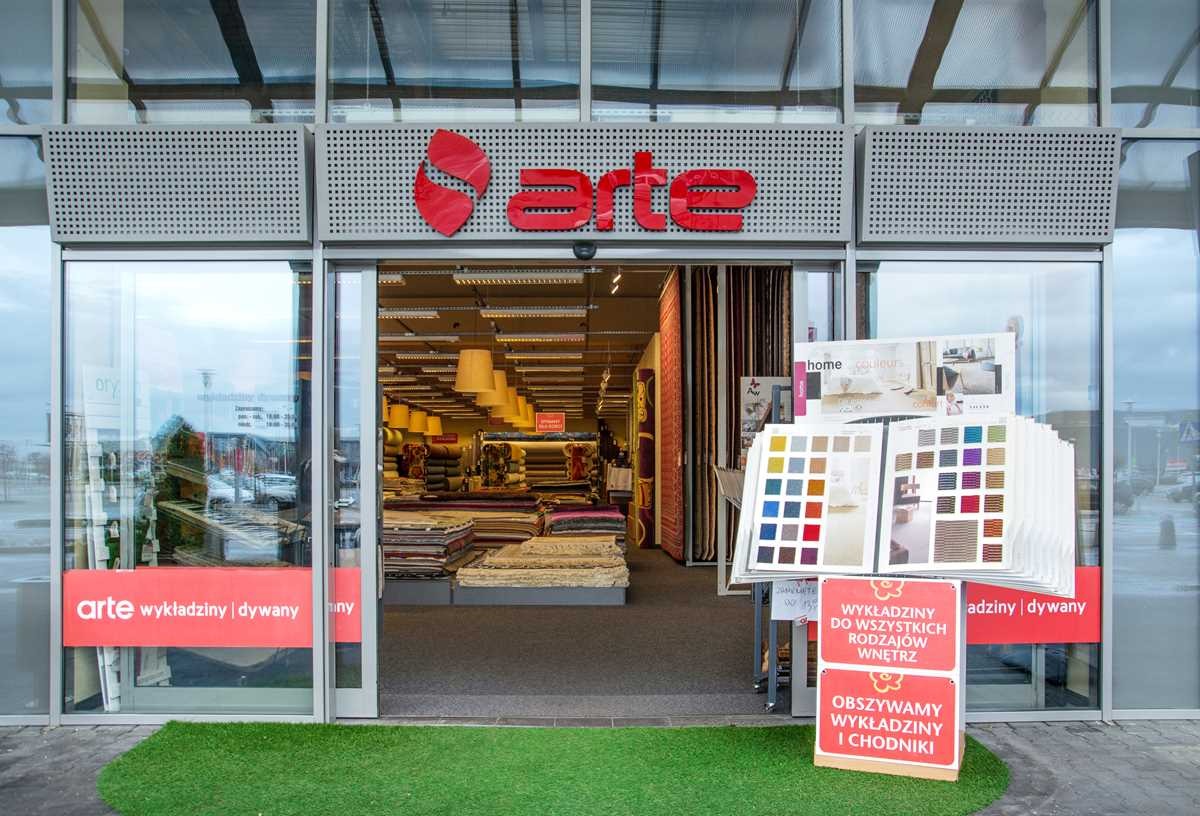 Wejście do sklepu z asortymentem podłogowym, widoczne ekspozycje z próbkami kolorów i faktur dywanów oraz wykładzin, logo 'arte' nad wejściem.