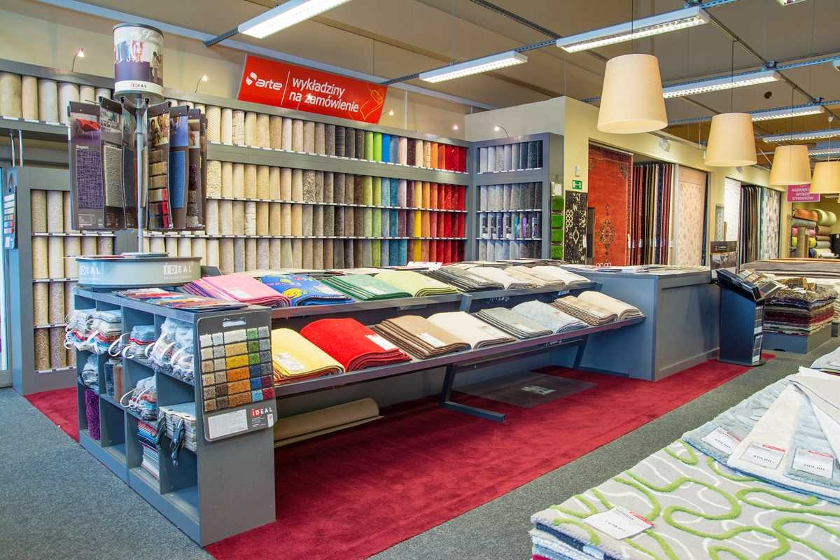 Ekspozycja w sklepie z różnorodnymi próbkami dywanów i wykładzin, ułożonymi kolorystycznie na półkach i prezentowanych na stojakach, z czerwonym dywanem na podłodze.