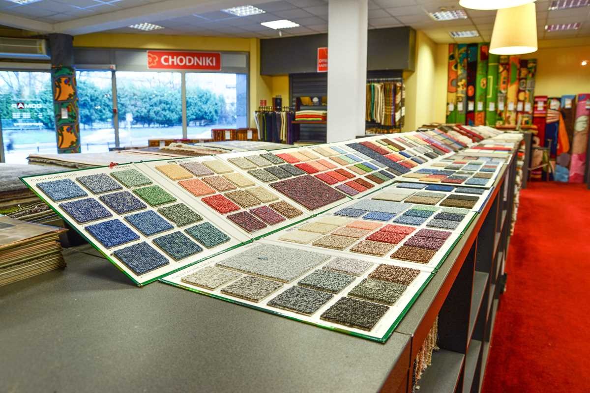 Ekspozycja próbek dywanowych w sklepie, widoczne różne kolory i tekstury, w tle czerwony dywan i regały z dywanami.