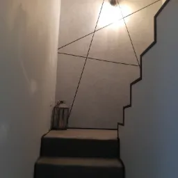 Schody z szarą wykładziną w Ostrołęce, ściana z dekoracyjną strukturą i geometrycznym wzorem z czarnych linii, lampion dekoracyjny u dołu schodów.